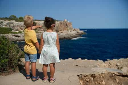 Dos niños cogidos de la mano y mirando el mar en una costa rocosa